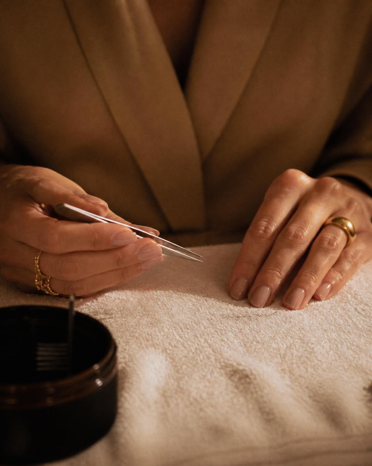 A lash artist's hands holding fine tweezers above a cream towel